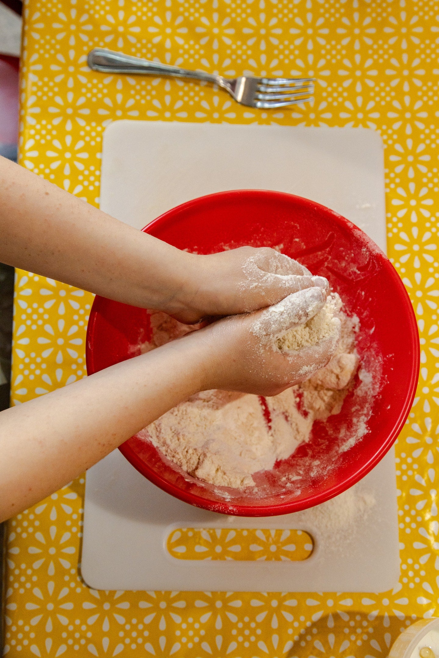 Person kneading dough in a red bowl on a yellow tablecloth with a fork.