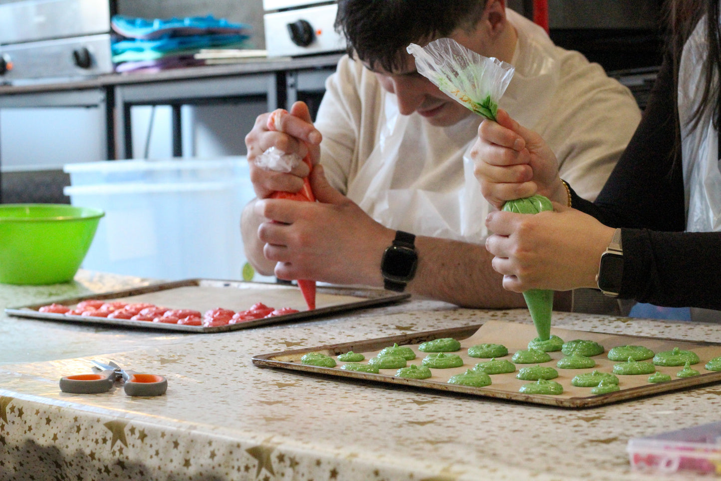 Two people pipe out their macarons onto baking sheets. The macarons on the left are bright pink, and the ones on the right are green.