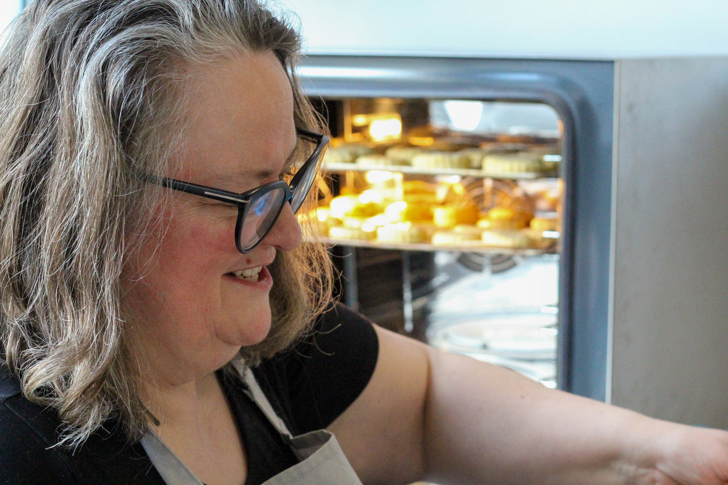 Course leader Joanna smiles a big smile in the foreground while scones bake in the oven behind her