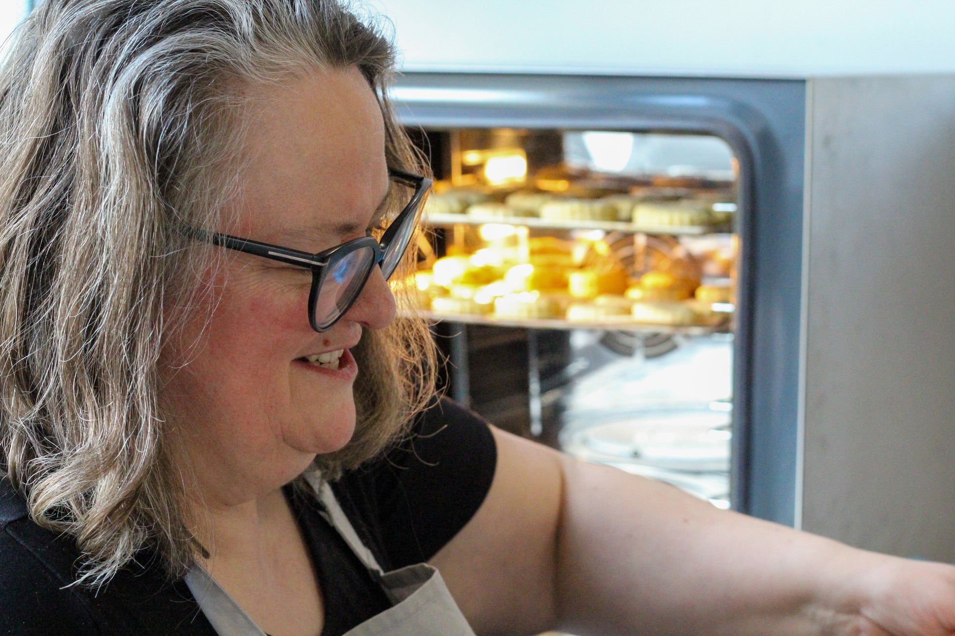 Course leader Joanna smiles a big smile in the foreground while scones bake in the oven behind her