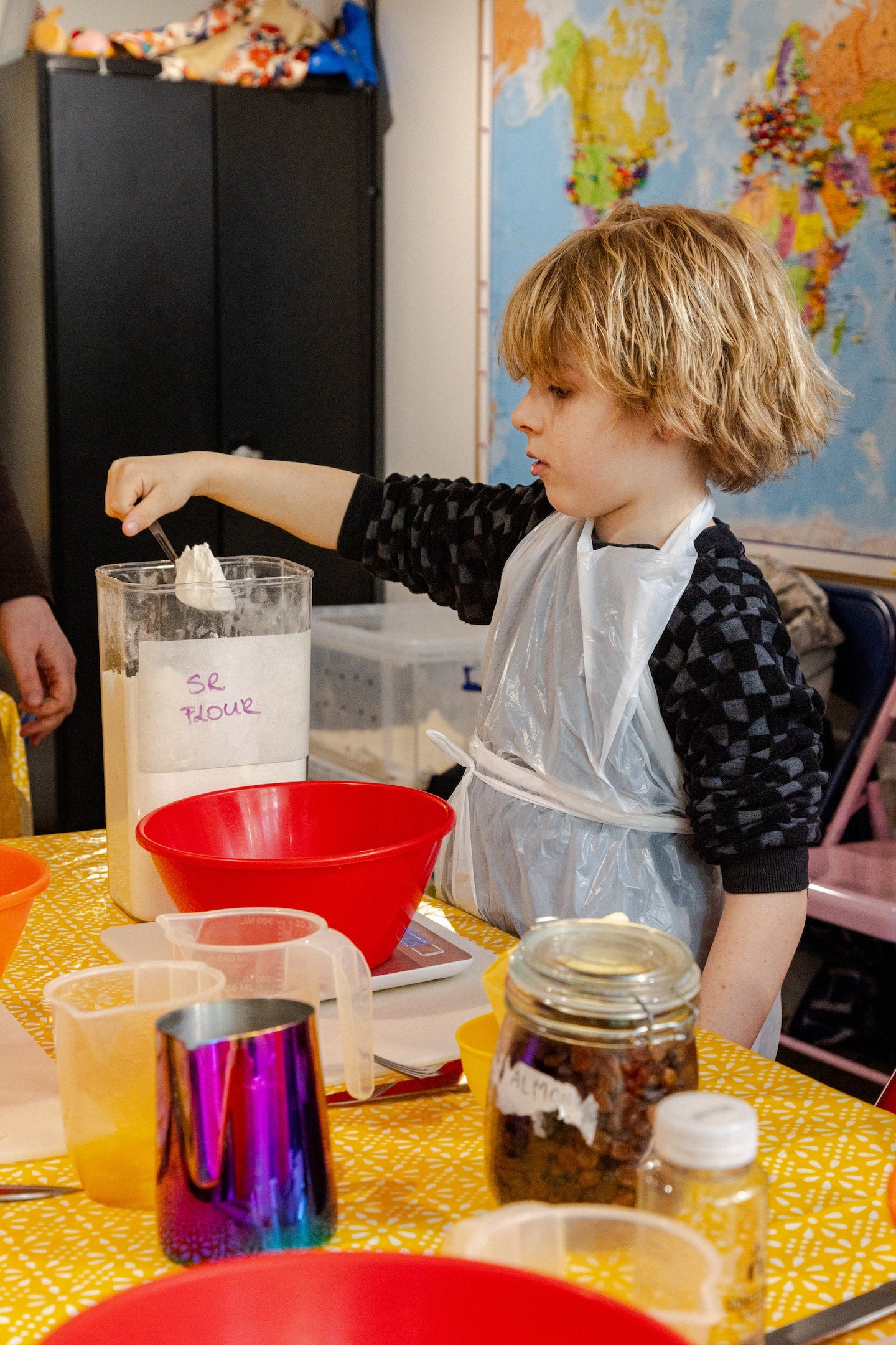 A child measures flour from a seethrough container into a mixing bowl