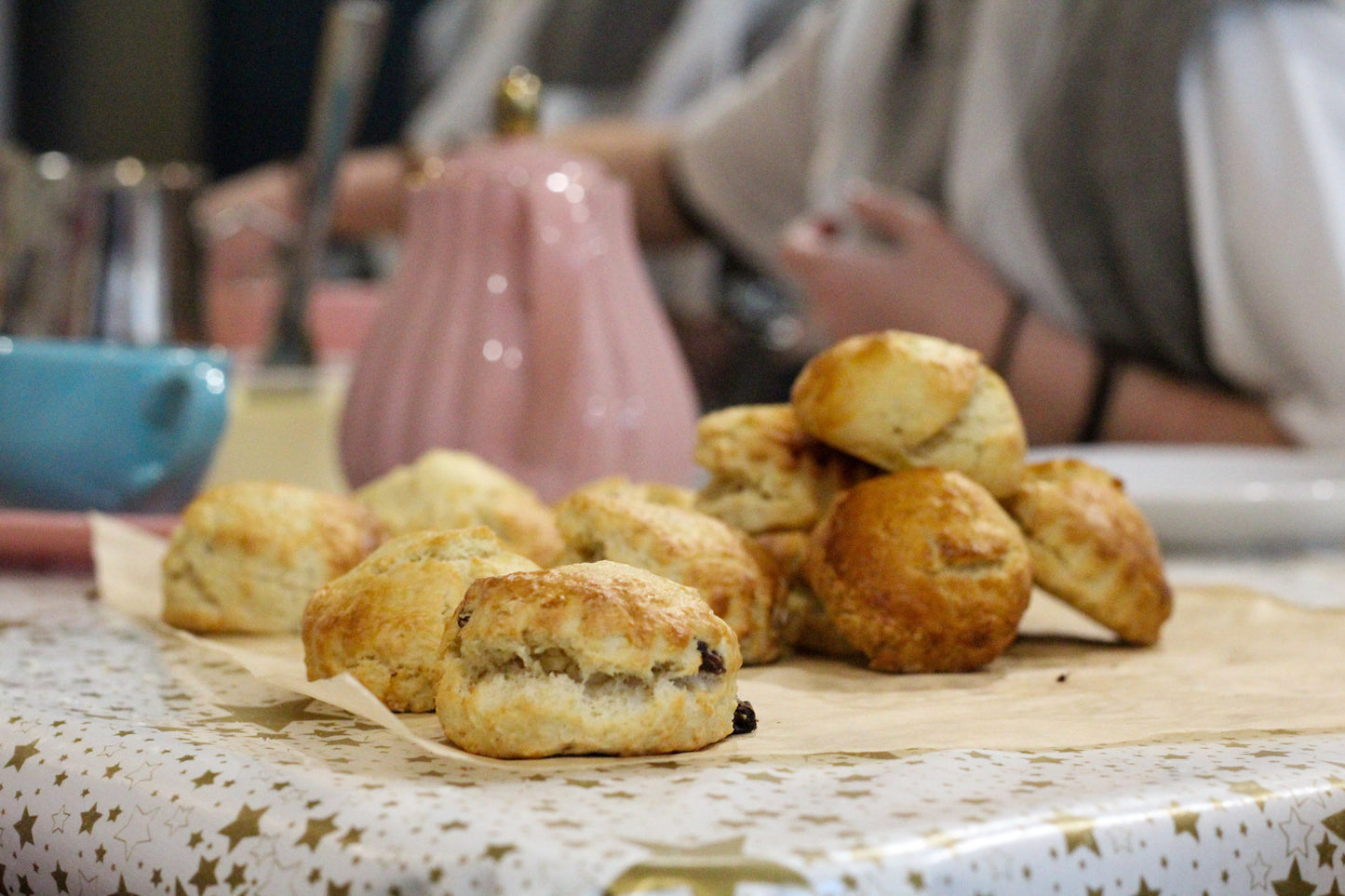 Scones are piled up in the foreground while a tea set is out of focus in the background