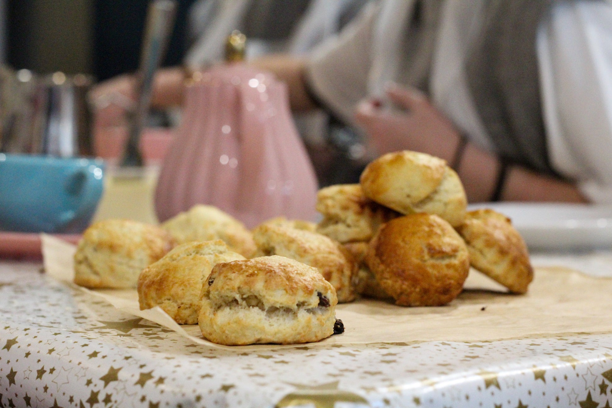 Scones are piled up in the foreground while a tea set is out of focus in the background