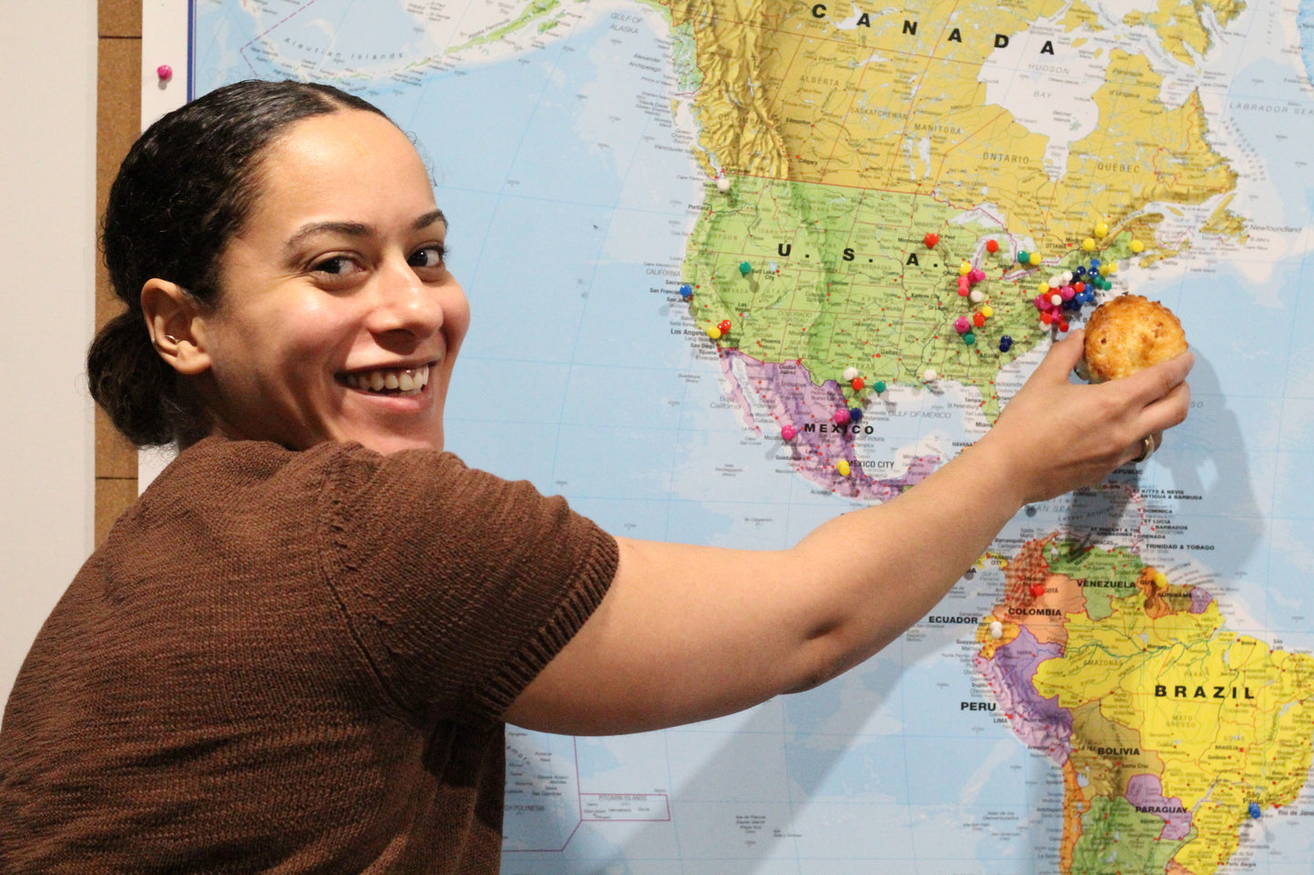 A smiling woman holds the scone she's just made up to a map next to where she's from - the East Coast of the United States.