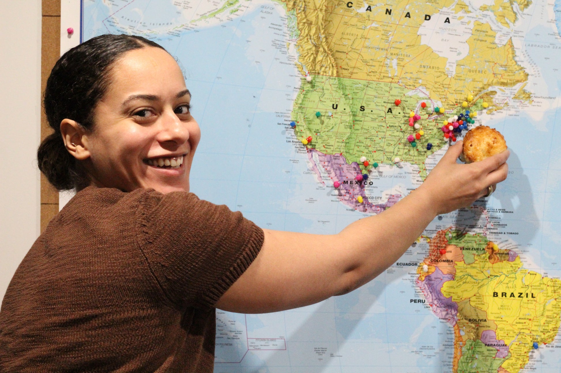 A smiling woman holds the scone she's just made up to a map next to where she's from - the East Coast of the United States.