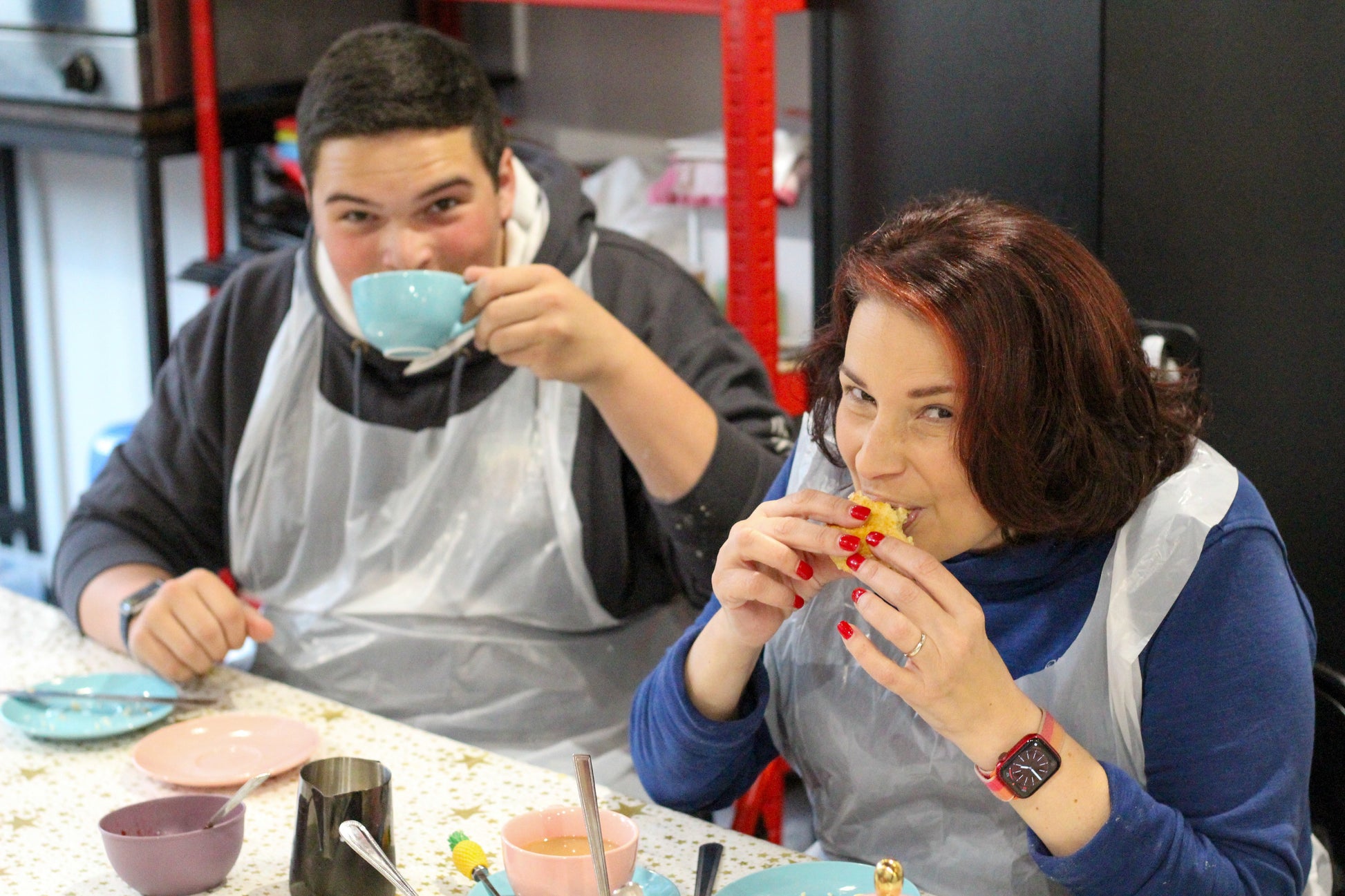 Two people look at camera. The man on the left is sipping from a blue cup while the other eats a scone with a mischievous look on her face.