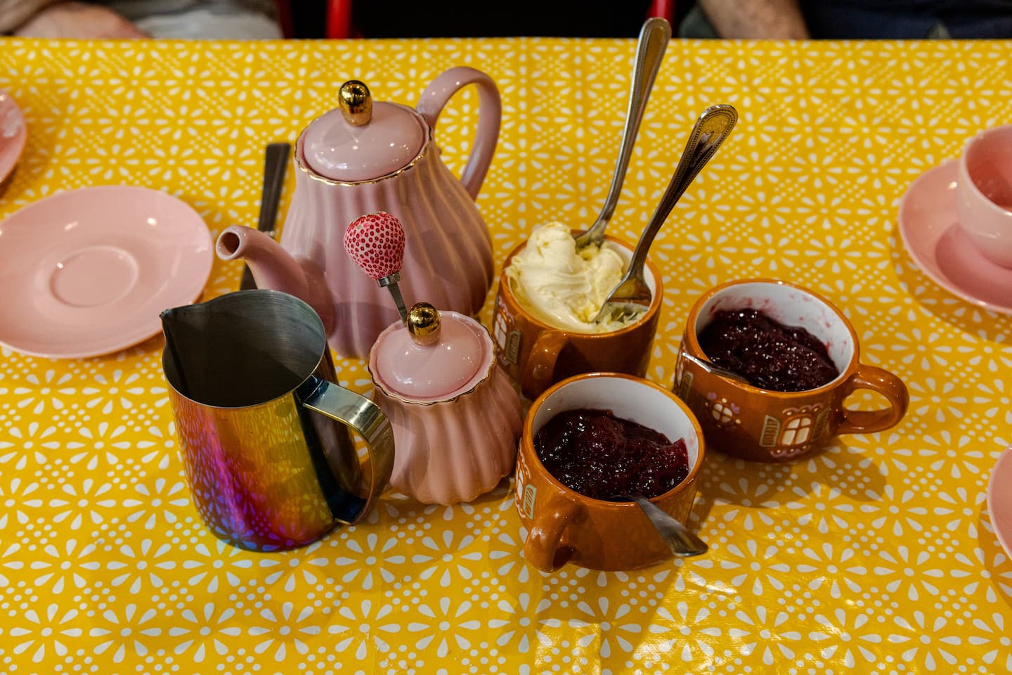 Tea set with pink teapot, mugs, and spoons on a yellow tablecloth. Jam and clotted cream sit in small pretty cups ready for spreading onto the freshly-baked scones.