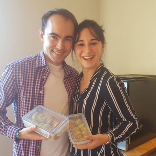 A man and a woman smile at the camera as they show off their freshly-made macarons in tupperware, ready to take home.