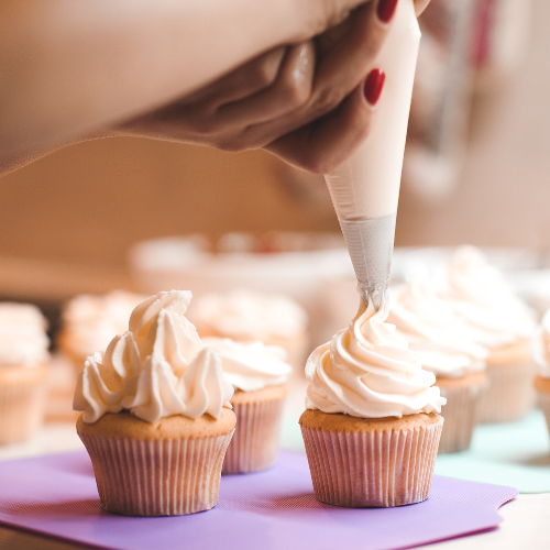 Learn to bake and decorate Cupcakes lilke a professional at one of our cupcake virtual workshops. Images shows someone carefully icing a cupcake with white icing