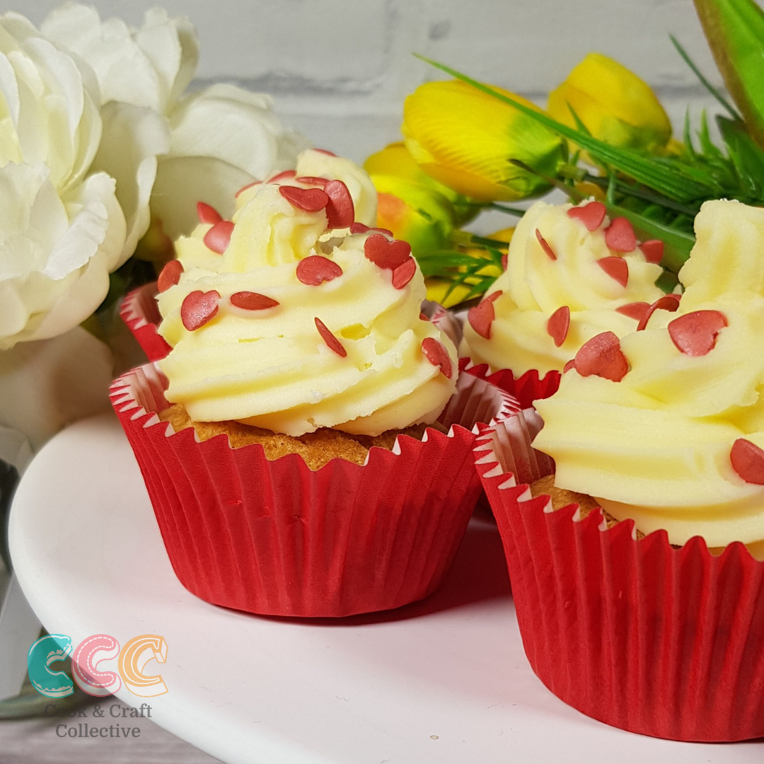Close up of some cupcakes with red cases, the white icing has pink love hearts on, in the background are some flowers for the added romantic feel
